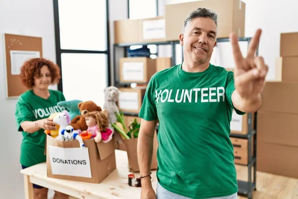 Middle age man wearing volunteer t shirt at donations stand smiling looking to the camera showing fingers doing victory sign. number two.