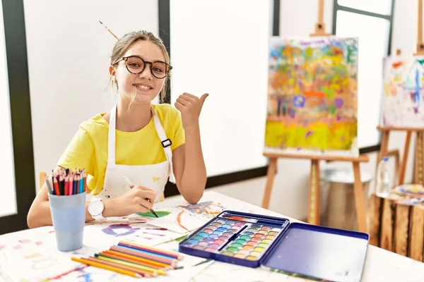 艺术工作室里的年轻的黑发少女 手举着大拇指指向后面 带着自信的微笑 — 图库照片
