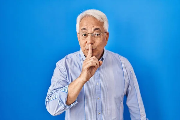 Hispanic senior man wearing glasses asking to be quiet with finger on lips. silence and secret concept.