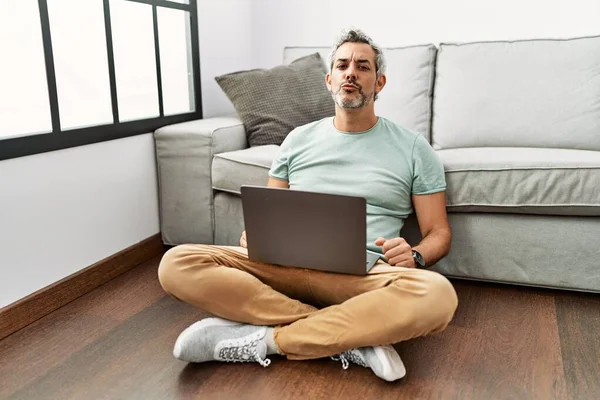 Middle age hispanic man using laptop sitting on the floor at the living room looking at the camera blowing a kiss on air being lovely and sexy. love expression.