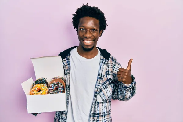 年轻的非洲裔美国男人拿着美味的彩色甜甜圈盒 开心而积极地微笑着 大拇指向上做得出色 签了字 — 图库照片