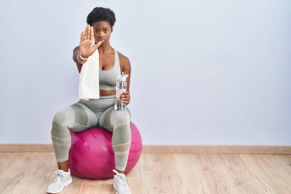 African american woman wearing sportswear sitting on pilates ball doing stop sing with palm of the hand. warning expression with negative and serious gesture on the face.