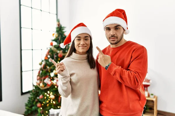 Young Hispanic Couple Standing Christmas Tree Pointing Hand Finger Side — Stok fotoğraf