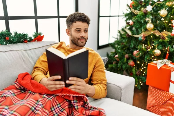 Young arab man reading book lying on the sofa by christmas tree at home.