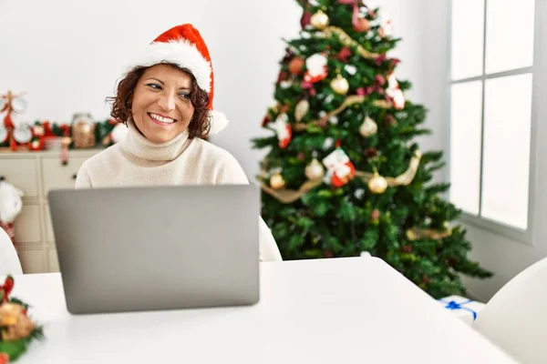 中世のヒスパニック系の女性のノートパソコンのクリスマスツリーの距離の顔に笑顔で横に見て座って使用して 自然な表現 自信を持って — ストック写真