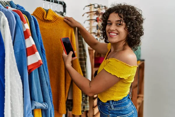 Young latin customer woman smiling happy choosing clothes using smartphone at clothing store.