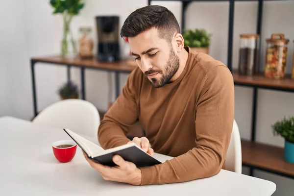 Young Hispanic Man Reading Book Drinking Coffee Sitting Table Home — Zdjęcie stockowe