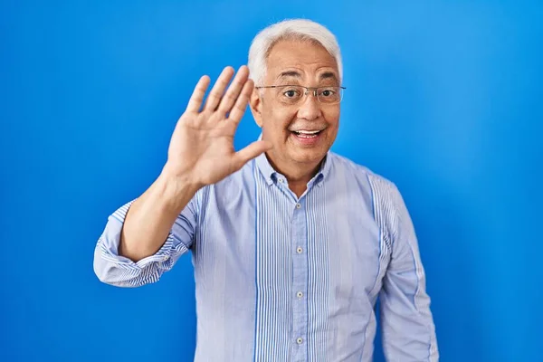 Hispanic senior man wearing glasses waiving saying hello happy and smiling, friendly welcome gesture
