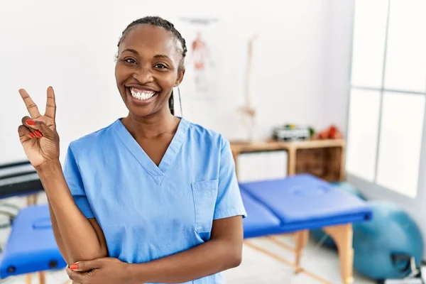 Black woman with braids working at pain recovery clinic smiling with happy face winking at the camera doing victory sign with fingers. number two.