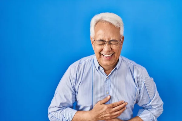 Hispanic senior man wearing glasses smiling and laughing hard out loud because funny crazy joke with hands on body.