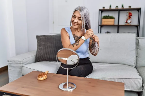 Middle age grey-haired woman combing hair with hands sitting on sofa at home