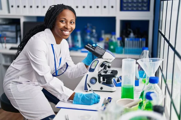 African American Woman Wearing Scientist Uniform Writing Clipboard Using Microscope — Stockfoto