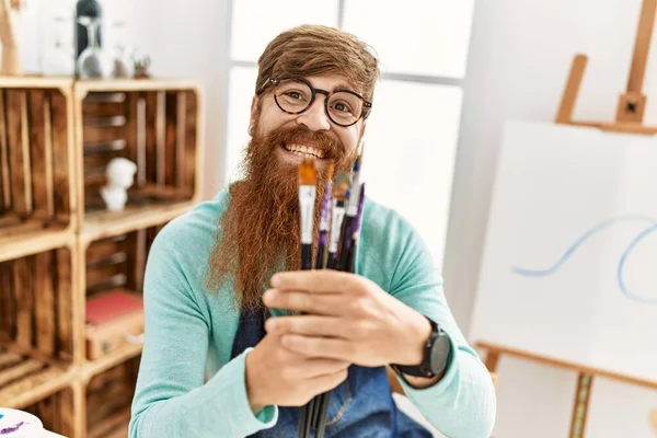 Young redhead man smiling confident holding paintbrushes at art studio