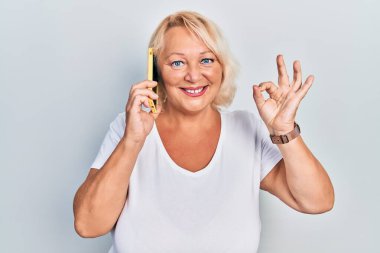 Middle age blonde woman having conversation talking on the smartphone doing ok sign with fingers, smiling friendly gesturing excellent symbol 