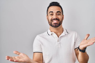 Beyaz arka plan üzerine gündelik giysiler giymiş sakallı genç İspanyol adam güler yüzlü, kolları açık, dostça karşılama, olumlu ve kendinden emin selamlar. 