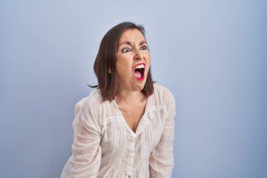 Middle age hispanic woman standing over blue background angry and mad screaming frustrated and furious, shouting with anger. rage and aggressive concept. 