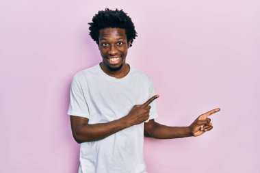 Beyaz bir tişört giymiş genç Afro-Amerikalı adam gülümsüyor ve kameraya bakıyor iki eliyle ve parmaklarıyla kenarı işaret ediyor.. 