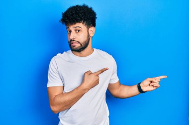 Sakallı, beyaz tişörtlü genç bir Arap. Endişeli ve gergin. Her iki eli de endişeli ve şaşırmış. 