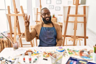 Sanat stüdyosundaki Afrikalı Amerikalı sanatçı kendini beğenmiş ve mutlu bir şekilde gülümserken üç numarayı gösterip işaret ediyor.. 