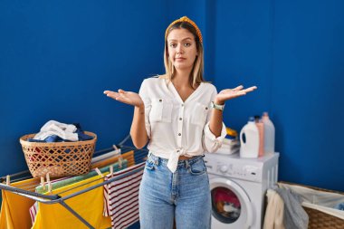 Çamaşır odasında elleri ve kolları havada, kafası karışmış genç sarışın bir kadın. şüphe kavramı. 