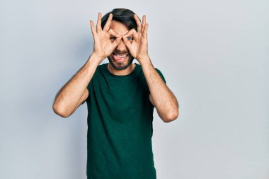 Latin asıllı genç bir adam, günlük beyaz tişört giyiyor dürbün gibi jest yapıyor, dili dışarı çıkarıyor, gözleri parmaklarından bakıyor. çılgın ifade. 