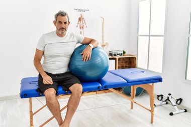 Orta yaşlı İspanyol bir adam ağrı kesici kliniğinde pilates topunu kuşkucu ve gergin tutuyor, sorun yüzünden surat asıyor. negatif kişi. 