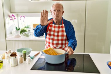 Ev mutfağında spagetti pişiren, gri saçlı son sınıf öğrencisi ciddi ve kendinden emin bir ifadeyle dur işareti yapıyor. 