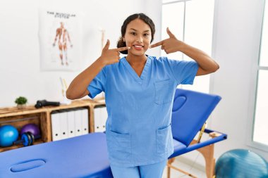 Güzel İspanyol fizyoterapist kadın ağrıyı dindirme kliniğinde gülümsüyor ve parmaklarıyla ve ağzıyla işaret ediyor. diş sağlığı kavramı. 