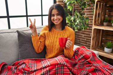 Kanepede oturan, gülümseyen, kameraya göz kırpan ve parmaklarıyla zafer işareti yapan İspanyol bir kadın. İki numara.. 