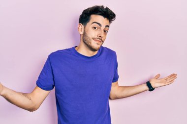 Gündelik tişört giyen, elleri ve kolları havada, kafası karışık, genç İspanyol bir adam. şüphe kavramı. 