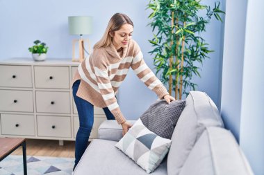 Gülümseyen genç bir kadın evdeki kanepeye yastık koyuyor.