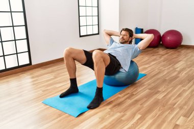 Orta yaşlı, beyaz, gülümseyen, kendine güvenen bir adam spor merkezinde formda top kullanıyor.