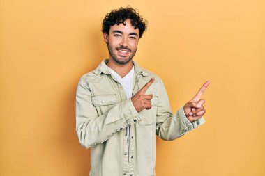 Gündelik kıyafetler giyen genç İspanyol adam gülümseyip kameraya bakıyor ve iki eliyle ve parmaklarıyla kenarı işaret ediyor.. 