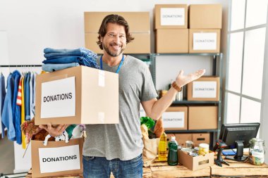 Handsome middle age man holding donations box for charity at volunteer stand smiling cheerful presenting and pointing with palm of hand looking at the camera. 