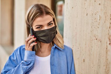 Coronavirus maskesi takan genç sarışın kız şehirdeki akıllı telefondan konuşuyor..
