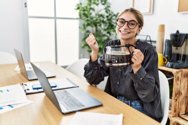 Ofiste çalışan genç sarışın kadın elinde kahve makinesiyle gururla bağırıyor, zaferi kutluyor ve kollarını kaldırarak çok heyecanlanıyor. 
