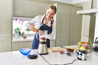 Genç sarışın kadın mutfaktaki miksere süt dökerek gülümsüyor.