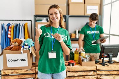 Young blonde girl wearing volunteer t shirt at donation stand pointing to the back behind with hand and thumbs up, smiling confident 