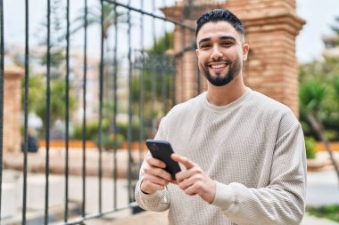 Genç Arap adam sokakta akıllı telefon kullanarak kendinden emin gülümsüyor.