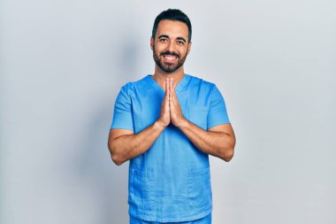 Yakışıklı, sakallı, mavi erkek hemşire üniforması giyen, elleriyle dua eden, affedilmek isteyen, kendinden emin, gülümseyen bir İspanyol.. 