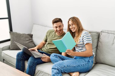 Genç İspanyol çift evde kanepede oturmuş dizüstü bilgisayar kullanıyor ve kitap okuyor..