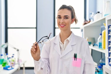 Young woman wearing scientist uniform holding glasses at laboratory