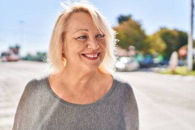 Orta yaşlı sarışın kadın sokakta kendinden emin bir şekilde gülümsüyor.