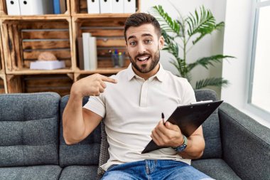 Psikoloji kliniğinde çalışan yakışıklı İspanyol bir adam yüzünde bir gülümsemeyle gururlu ve mutlu birini işaret ediyor.. 