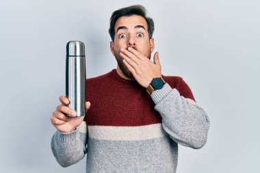 Sakallı, ağzı elle kaplanmış termo tutan yakışıklı, beyaz bir adam. Şok olmuş ve hata yapmaktan korkuyor. şaşırmış ifade 