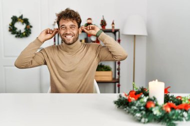 Masada sakallı, genç yakışıklı bir adam Noel süslemesinin yanında gülümsüyor kulakları parmaklarıyla çekiyor, komik bir jest. deneme sorunu 