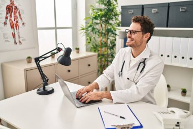 Young hispanic man wearing doctor uniform using laptop at clinic