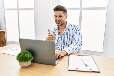 Young handsome man with beard working at the office using computer laptop doing happy thumbs up gesture with hand. approving expression looking at the camera showing success. 