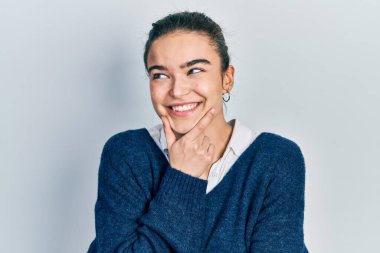 Elleri çenesinde gündelik giysiler giyen genç beyaz kız soru soruyor, düşünceli bir yüz ifadesi var. Düşünceli bir yüzle gülümsüyor. şüphe kavramı. 