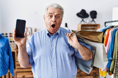 Elinde alışveriş poşetleri olan ve akıllı telefon ekranı gösteren yaşlı bir adam şaşkınlık ve şaşkınlık dolu bir ifade, korku ve heyecanlı bir yüz ifadesiyle korku içinde.. 